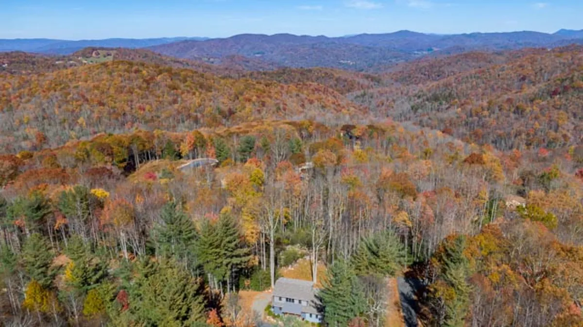 The house is shown surrounded by vibrant autumn foliage and an expansive mountain range in the background. The view encapsulates the essence of mountain walks, seasonal changes, and the serenity of nature accessible from the property.