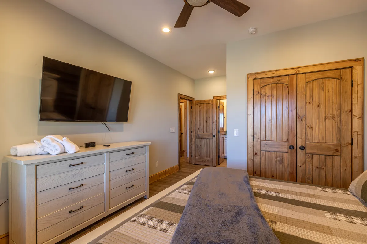 A welcoming bedroom with a large bed adorned with a decorative blanket, a wall-mounted television, and a wooden dresser. The room exudes a cozy and comfortable atmosphere, ideal for relaxation and privacy.