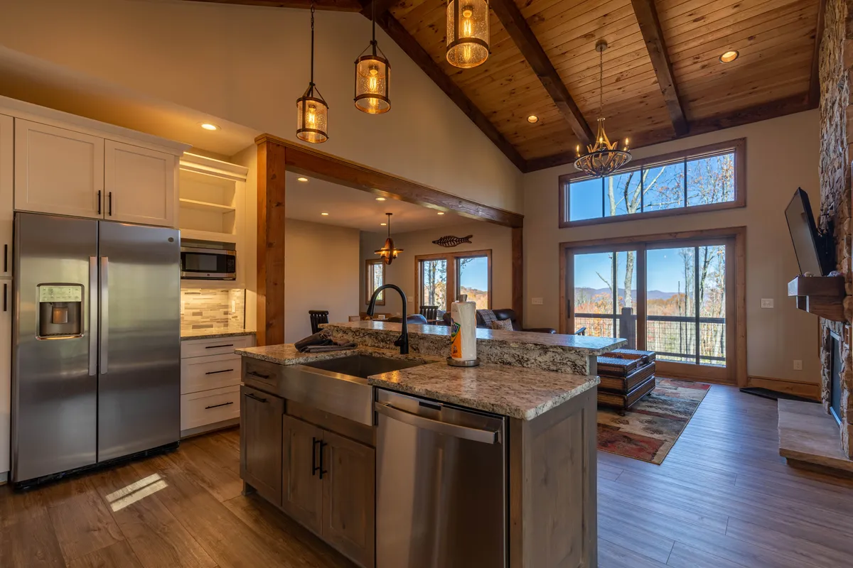 A bright, open-concept kitchen and living area with a sturdy kitchen island, wood-accented ceiling, and a stone fireplace. Large windows bathe the room in natural light and offer glimpses of the mountain scenery, suggesting a seamless transition between indoor comfort and the outdoor environment.