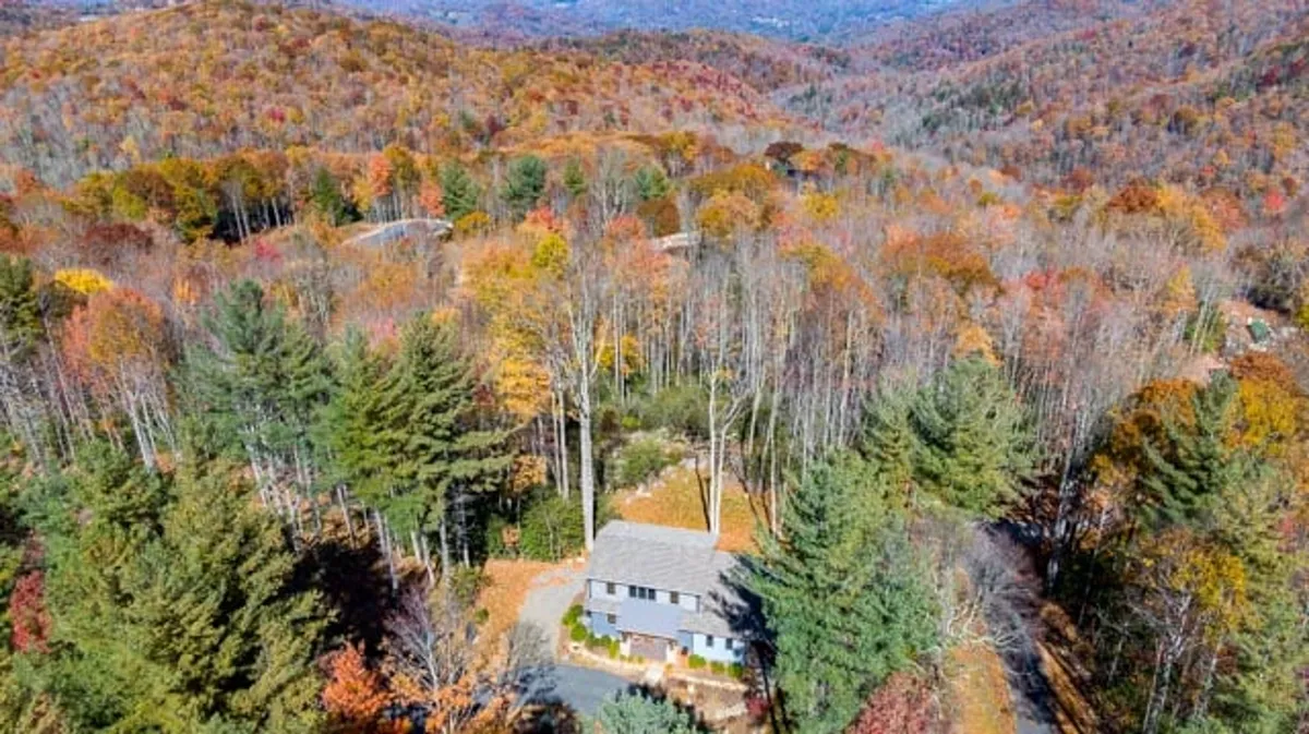 A serene exterior view of the house nestled among abundant autumn foliage and tall trees, with the surrounding mountain landscape hinting at nearby trails and outdoor adventures. The setting is tranquil, offering a perfect blend of natural beauty and privacy for those seeking work-life balance in the mountains.