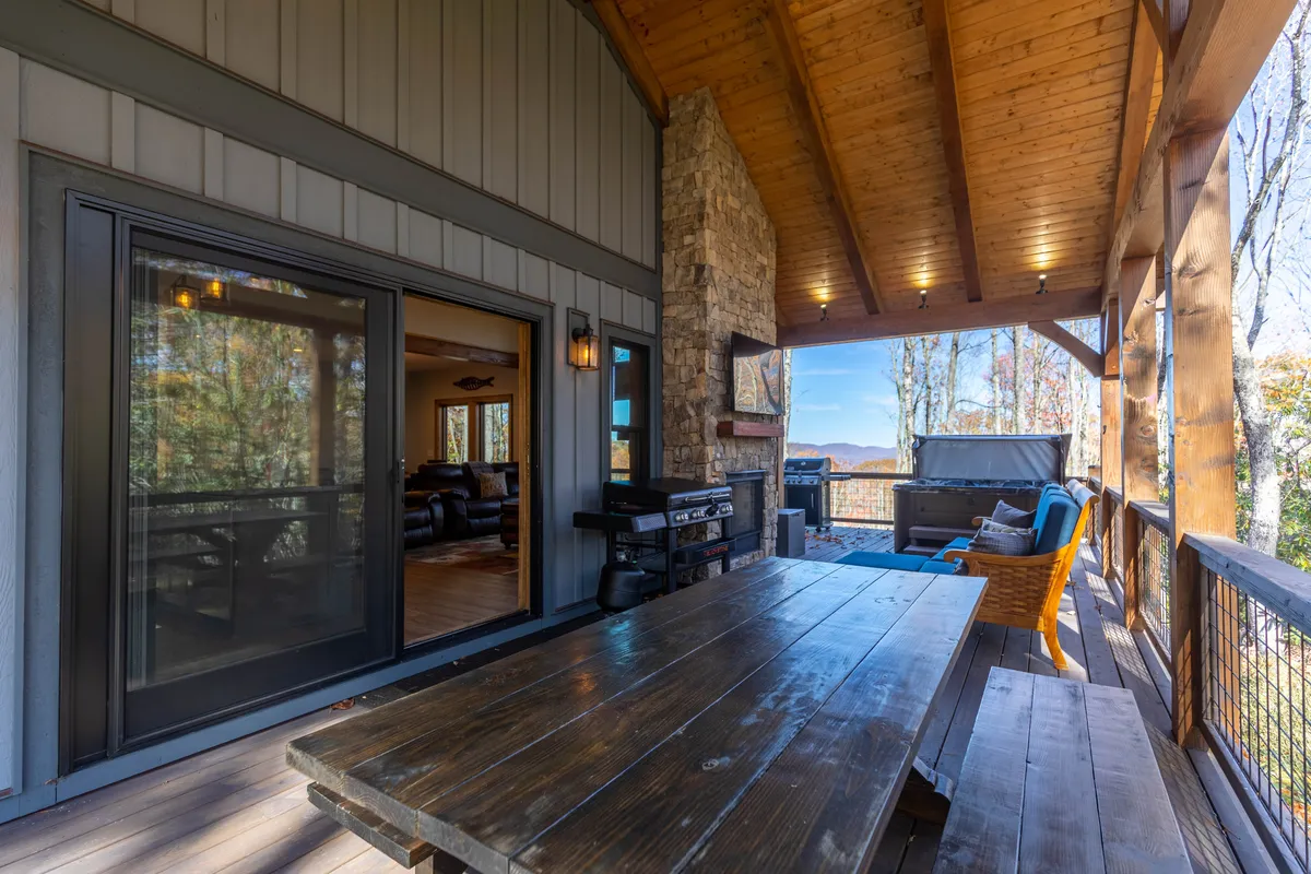 A large wooden dining table is set on the deck, complemented by a stone fireplace and comfortable seating. The panoramic view through the windows showcases the mountain scenery, suggesting possibilities for both alfresco dining and relaxed evenings with friends or family.