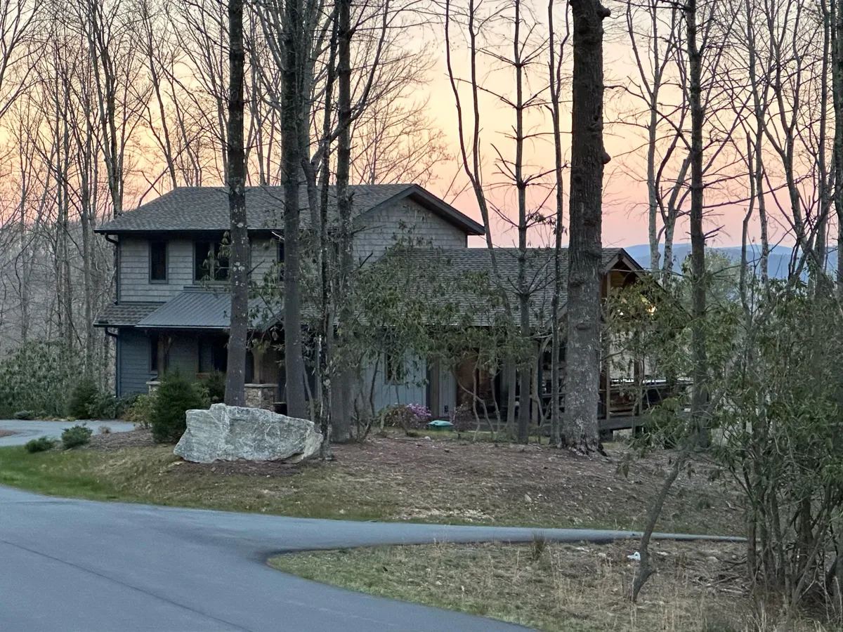 This image features the exterior of the house nestled among mature trees, with a prominent rock in the foreground. The porch invites relaxation and the natural setting highlights direct access to walking trails and outdoor space, ideal for pets and their owners.