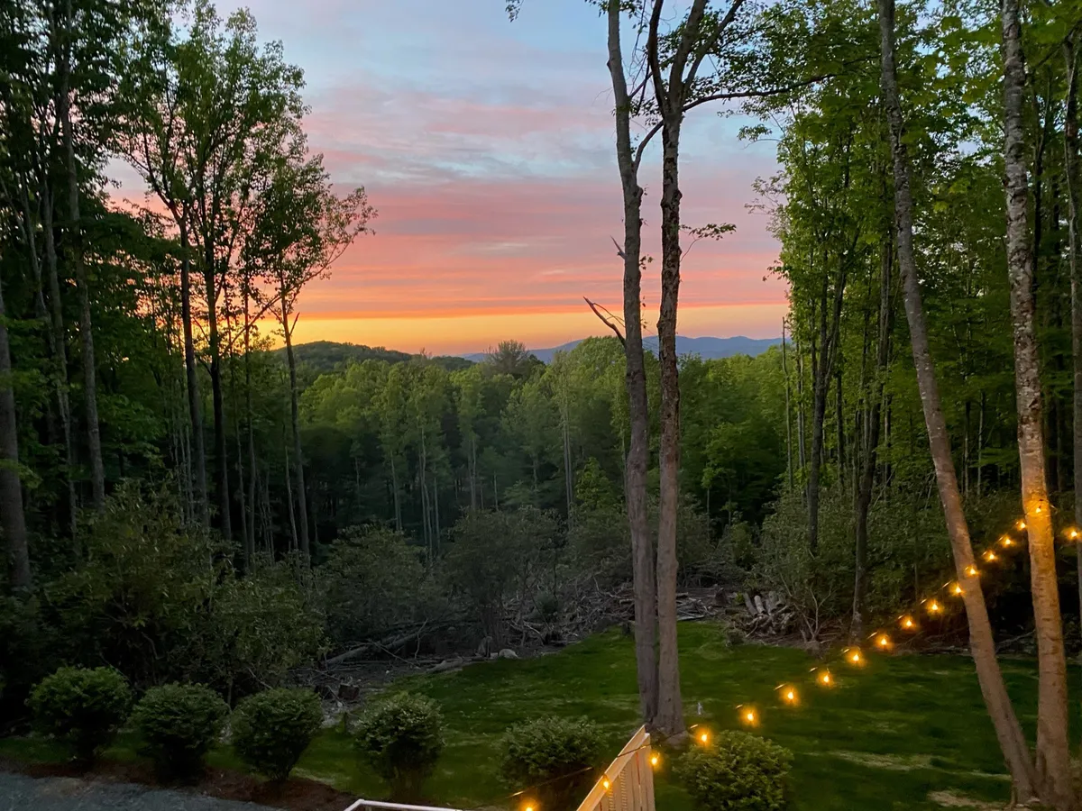 A sweeping vista of the mountain range at sunset, with dense trees below and soft string lights suggesting a welcoming outdoor space for relaxation or after-work gatherings. The inviting glow of twilight conveys tranquility and the unique experience of working in a mountain environment.