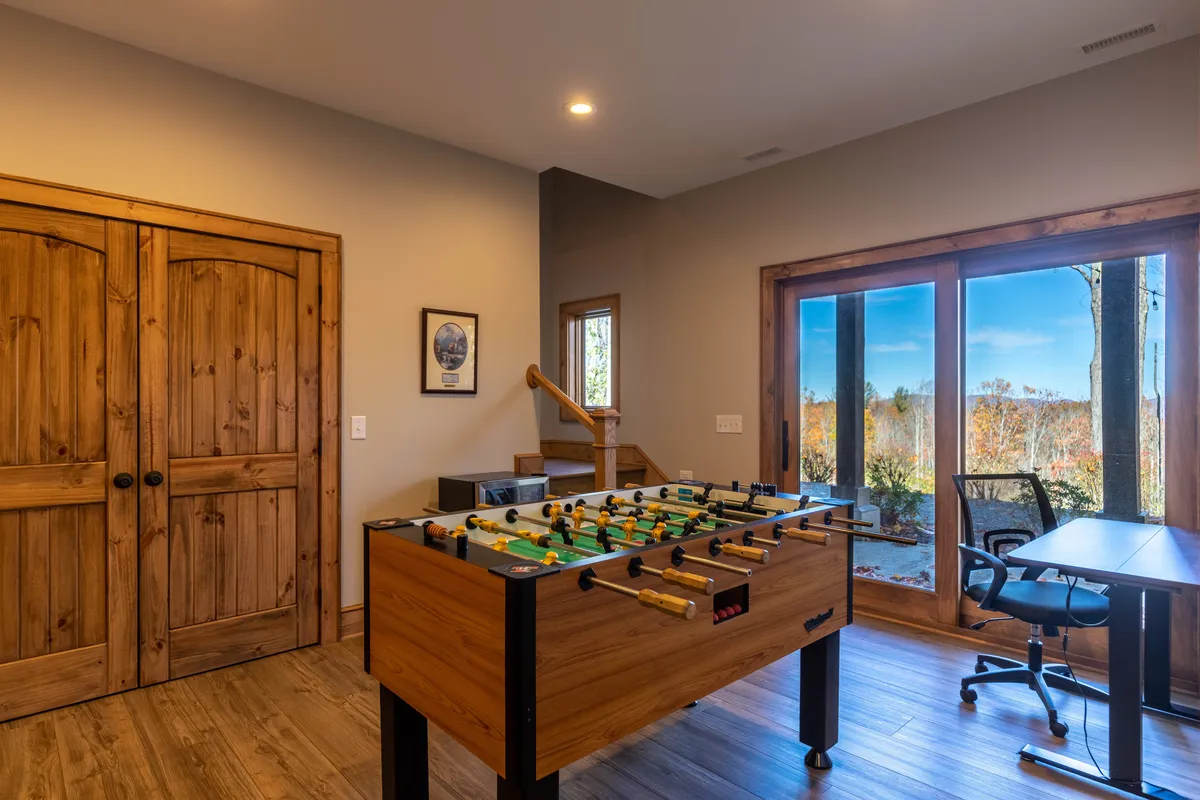 A playful interior with a foosball table set beside large windows and wooden doors opening to the outdoors. The space is designed for recreation and relaxation, inviting friendly competition and family game nights with views of the surrounding nature.