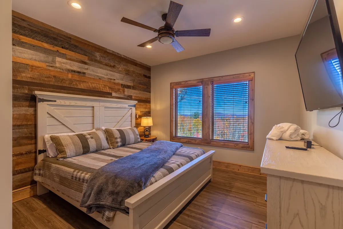 A tranquil bedroom featuring a plush bed against a wooden accent wall, a ceiling fan overhead, and a window that frames a scenic view. A flat-screen TV is mounted on the wall, and natural light fills the space, creating an inviting and restful atmosphere.