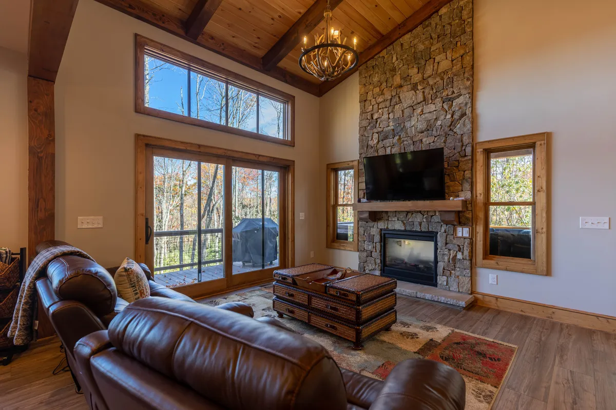 A spacious living area featuring a grand stone fireplace as the focal point, complemented by large windows that offer sweeping views of the outdoors. The inviting space includes a comfortable seating arrangement, a rustic coffee table, and a dramatic chandelier overhead. The environment exudes warmth and is perfect for gathering, relaxing after a day of adventure, or stargazing by the fire.