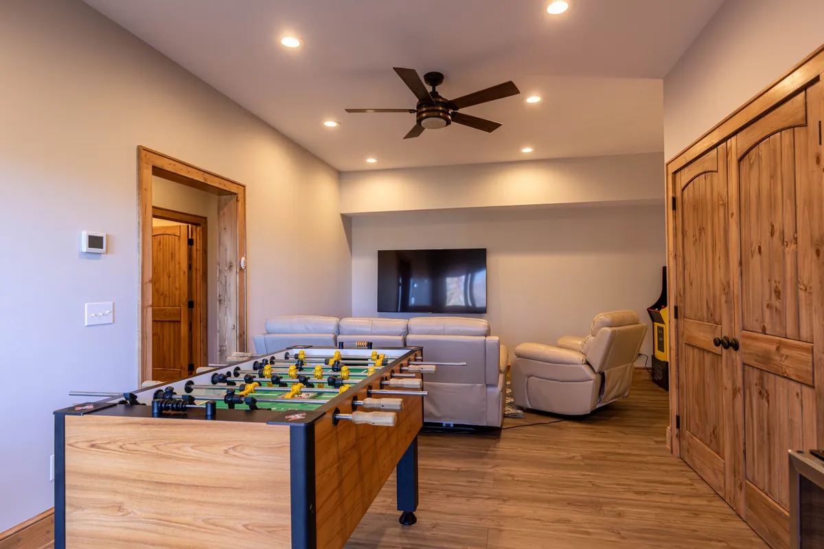 A basement game area with a foosball table positioned at the center, paired with reclining chairs and a TV set up for group gatherings. Wooden doors and neutral decor help create a casual, welcoming atmosphere for friendly competition and relaxation.