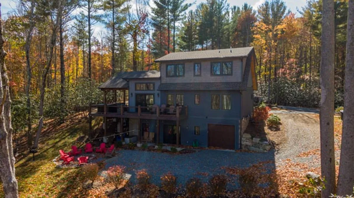 This image captures the inviting exterior of the two-story house, featuring a spacious deck with seating, red Adirondack chairs, and a backdrop of lush trees. The gravel driveway and welcoming outdoor spaces suggest ample room for guests and celebrations.