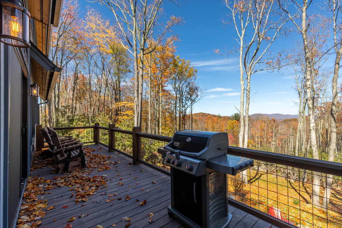 An inviting deck furnished with a grill and outdoor dining area, surrounded by vibrant fall foliage and offering an expansive mountain view in the distance. The cozy ambiance hints at tranquil mornings or intimate evenings spent outdoors, whether enjoying a meal, a glass of wine, or simply soaking up the natural beauty.