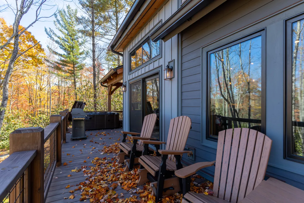 The image highlights a tranquil deck space featuring wooden chairs, a bubbling hot tub, and expansive views through large windows. The fall leaves add seasonal charm, and the space is well-suited for evening gatherings or stargazing sessions.