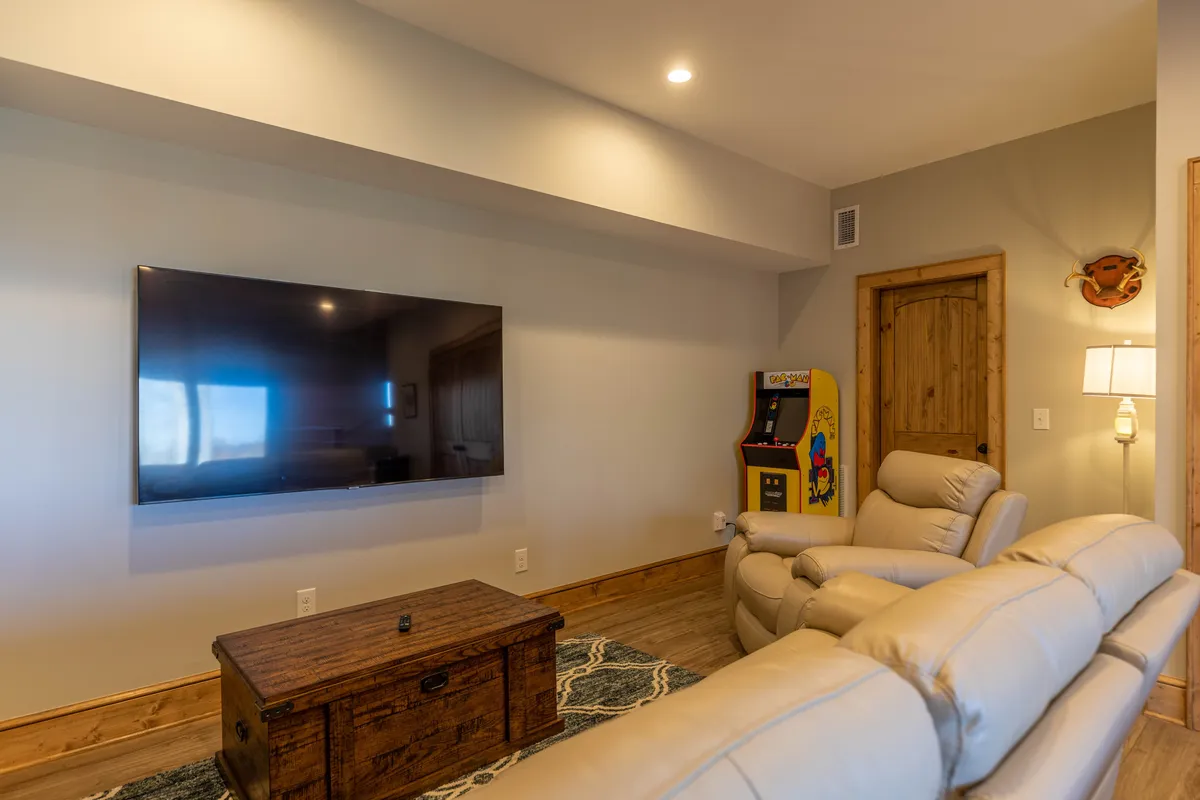 A spacious entertainment room featuring a large flat-screen TV mounted on the wall, an arcade game machine reminiscent of the classic PacMan, a reclining sofa set up for comfortable movie viewing, and a wooden coffee table. The space exudes a playful, inviting vibe and is clearly designed for both gaming and relaxing with friends or family.