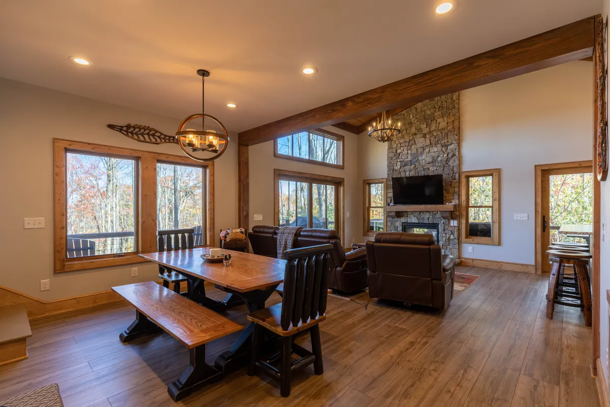 This image captures a wide view of the living and dining area, highlighting the spacious dining table, the impressive stone gas fireplace, large windows letting in natural light, and comfortable seating arranged for gathering. The ambiance suggests warmth and togetherness, with elements arranged for family meals, game nights, and relaxing by the fire.