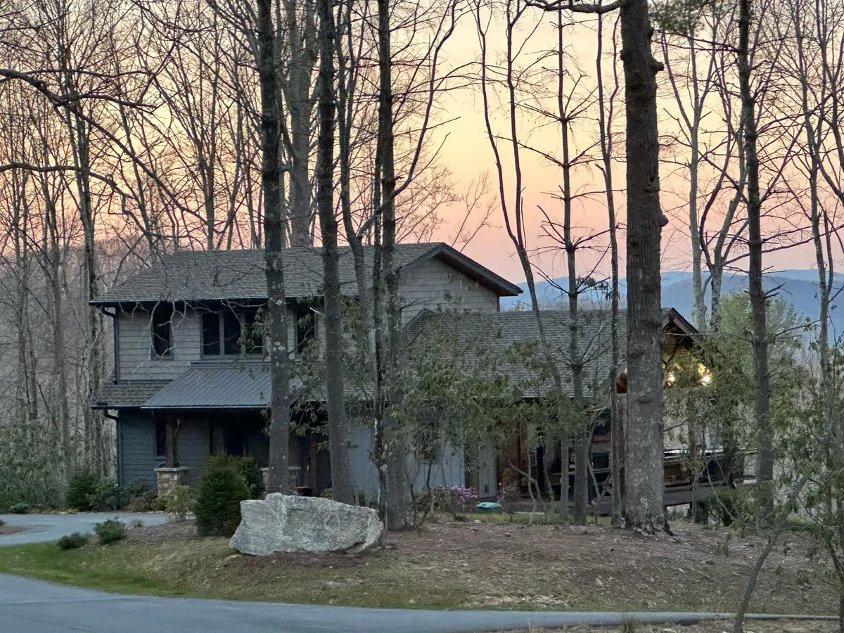 The image showcases the inviting exterior of a modern mountain home nestled among tall trees, with a stunning sunset painting the sky in warm hues. A spacious porch overlooks the natural landscape, hinting at opportunities for outdoor relaxation, while the secluded location promises peace and privacy.